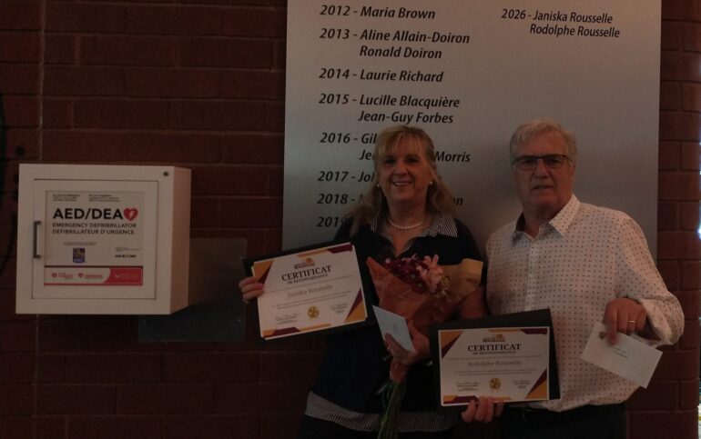 Photographie de Janiska et Rodolphe Rousselle posant devant la plaque des pionniers et bâtisseurs du Carrefour Beausoleil, fleurs et certificats en main.